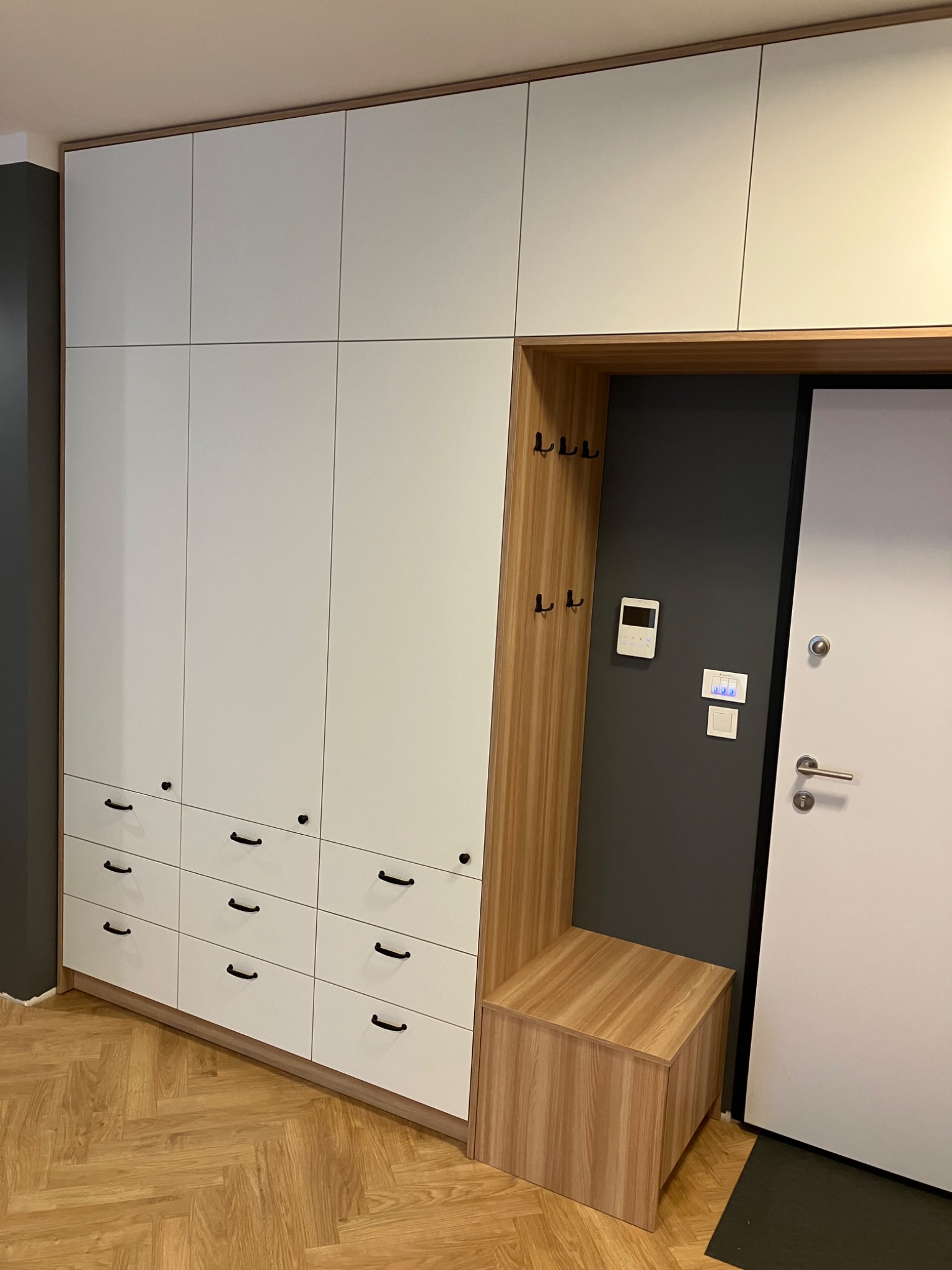 Biała szafa wnękowa z drewnianymi akcentami i siedziskiem przy wejściu, czarne uchwyty, parkiet w jodełkę, nowoczesny design.