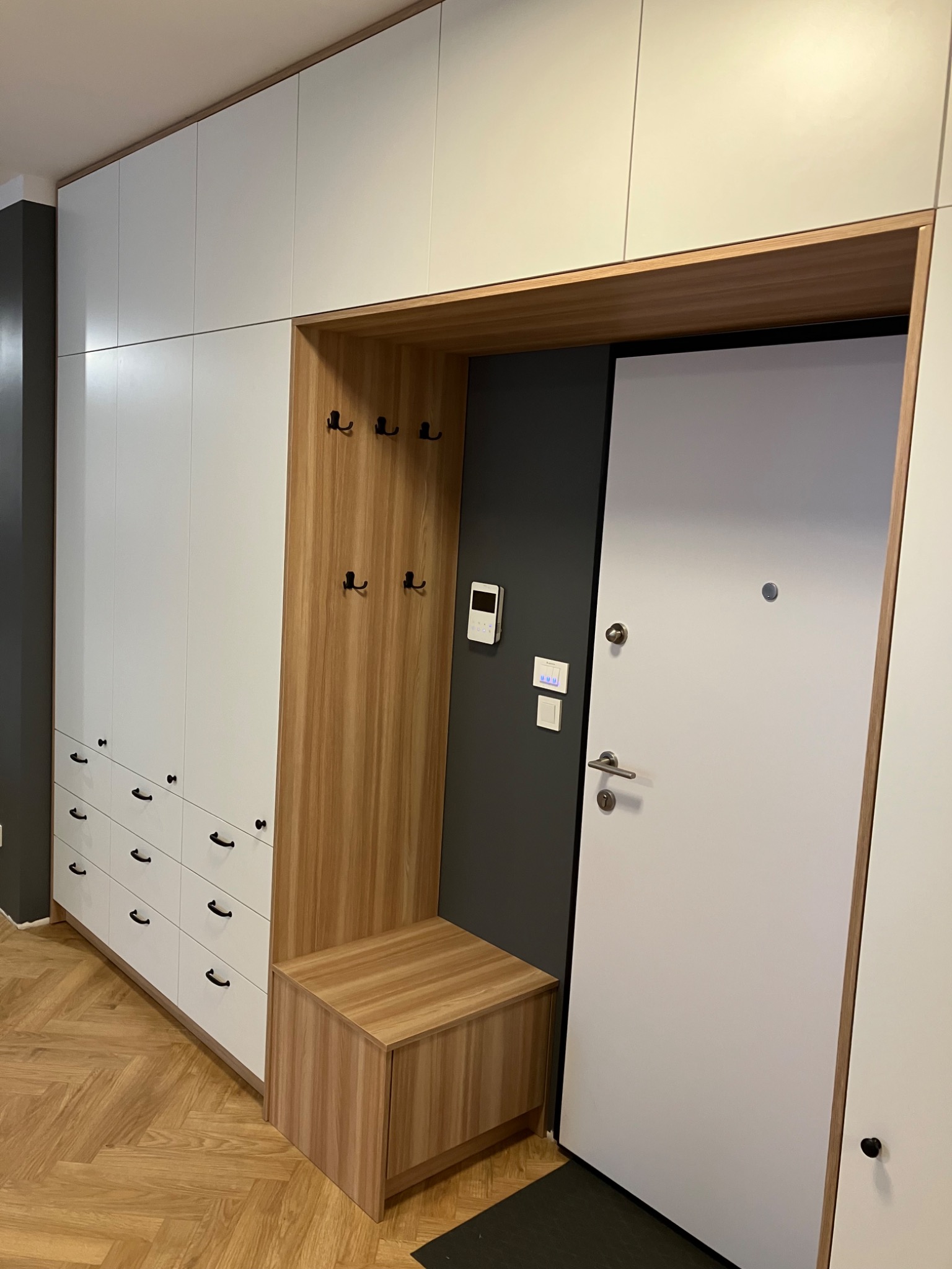 Biała szafa wnękowa z drewnianym siedziskiem i wieszakami w przedpokoju, obok drzwi wejściowych. Minimalistyczny design, jasne kolory, parkiet w jodełkę na podłodze.