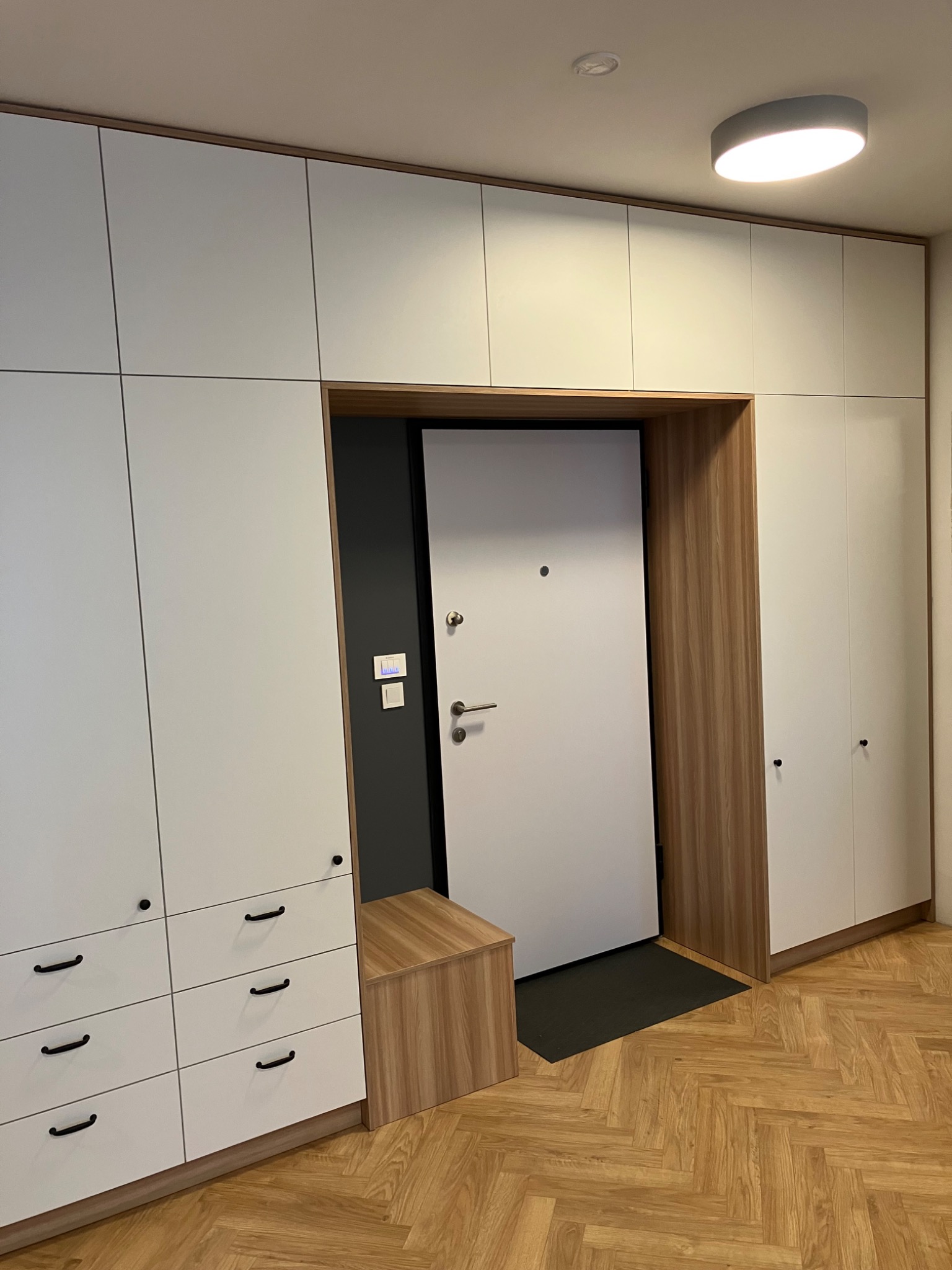 Białe szafy wnękowe z drewnianymi akcentami otaczają wejście z drzwiami. Widoczna drewniana podłoga w jodełkę i siedzisko. Minimalistyczny design, ciepłe oświetlenie.