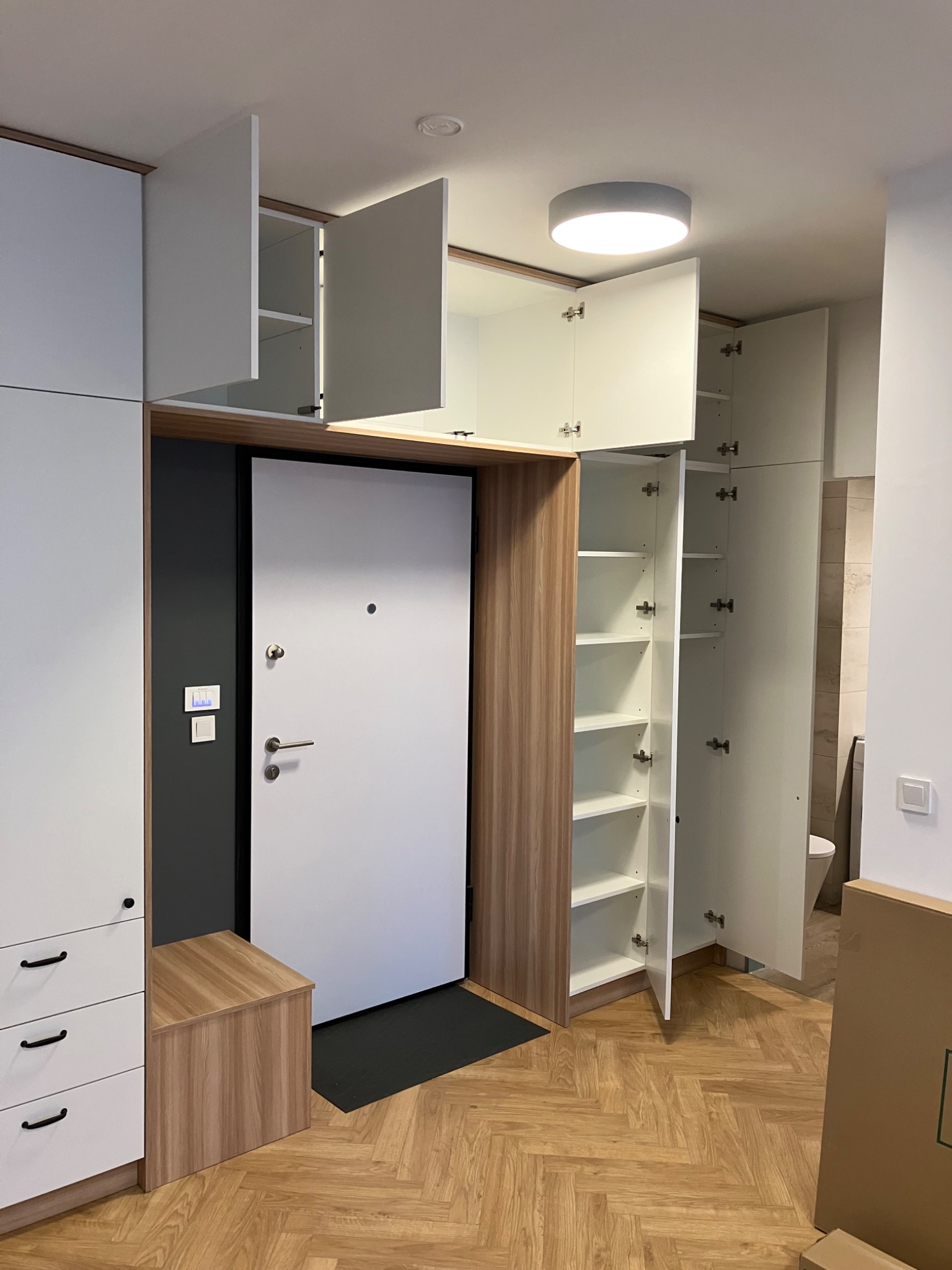 Biała szafa wnękowa z drewnianymi akcentami, wkomponowana we wnękę przy drzwiach wejściowych. Otwarta szafka z półkami i szuflady z czarnymi uchwytami.