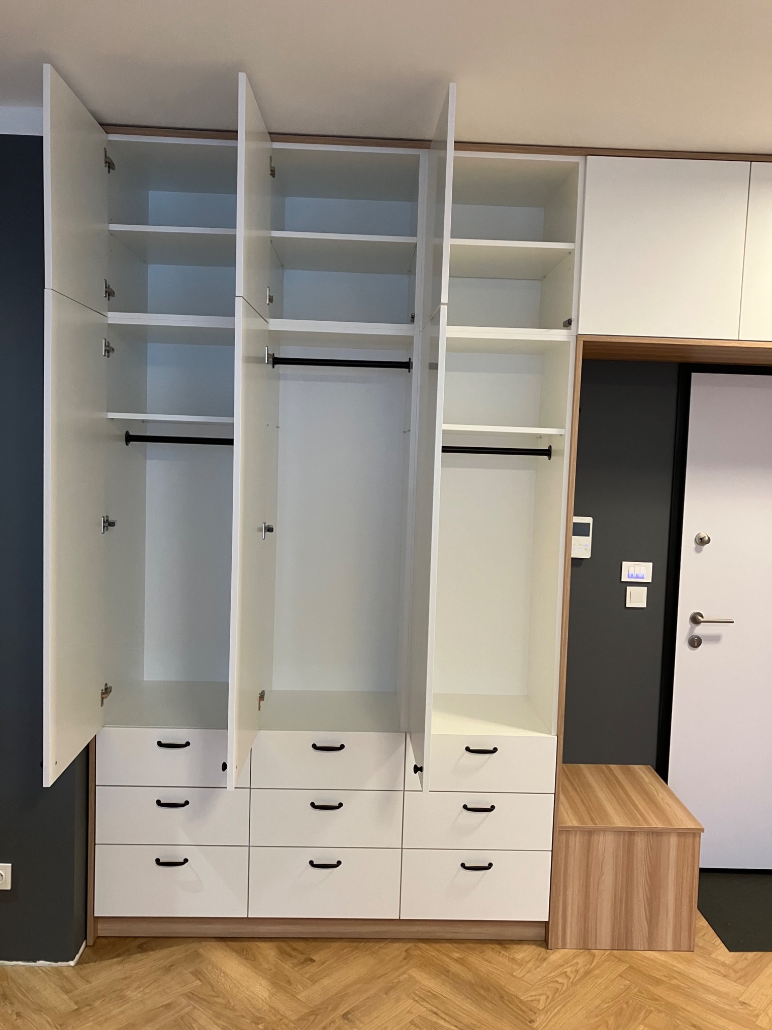 Biała szafa wnękowa z otwartymi drzwiami, widoczne półki i drążki na ubrania, dolne szuflady z czarnymi uchwytami, drewnopodobna podstawa i siedzisko obok.