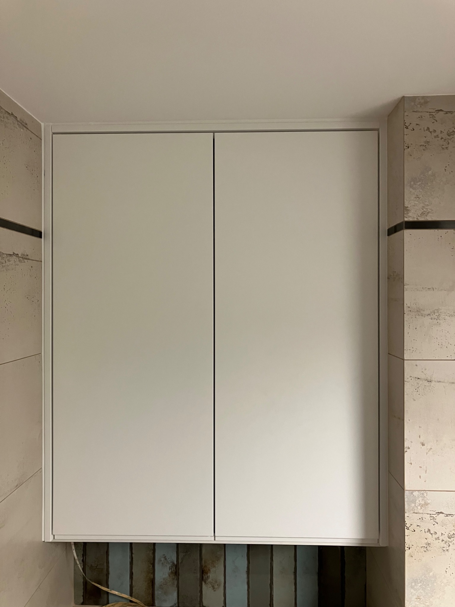 Biała szafka wnękowa z dwoma drzwiami, zamontowana w łazience z dekoracyjnymi płytkami. Widoczne przewody elektryczne u dołu szafki.