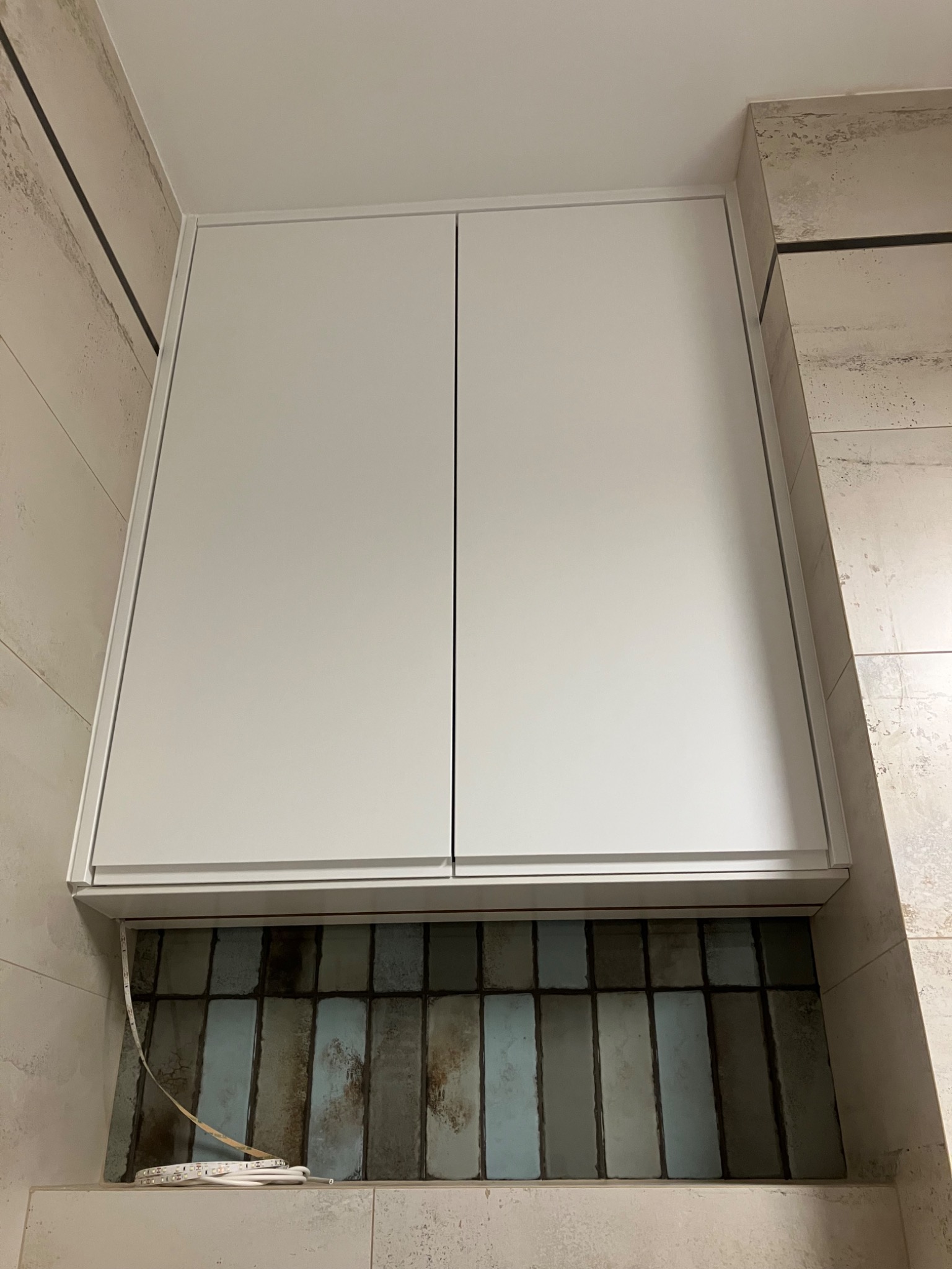 Biała szafka wisząca z dwoma drzwiczkami, zamontowana nad ścianą z dekoracyjnymi, wąskimi płytkami w odcieniach szarości i błękitu. Widoczna listwa LED.