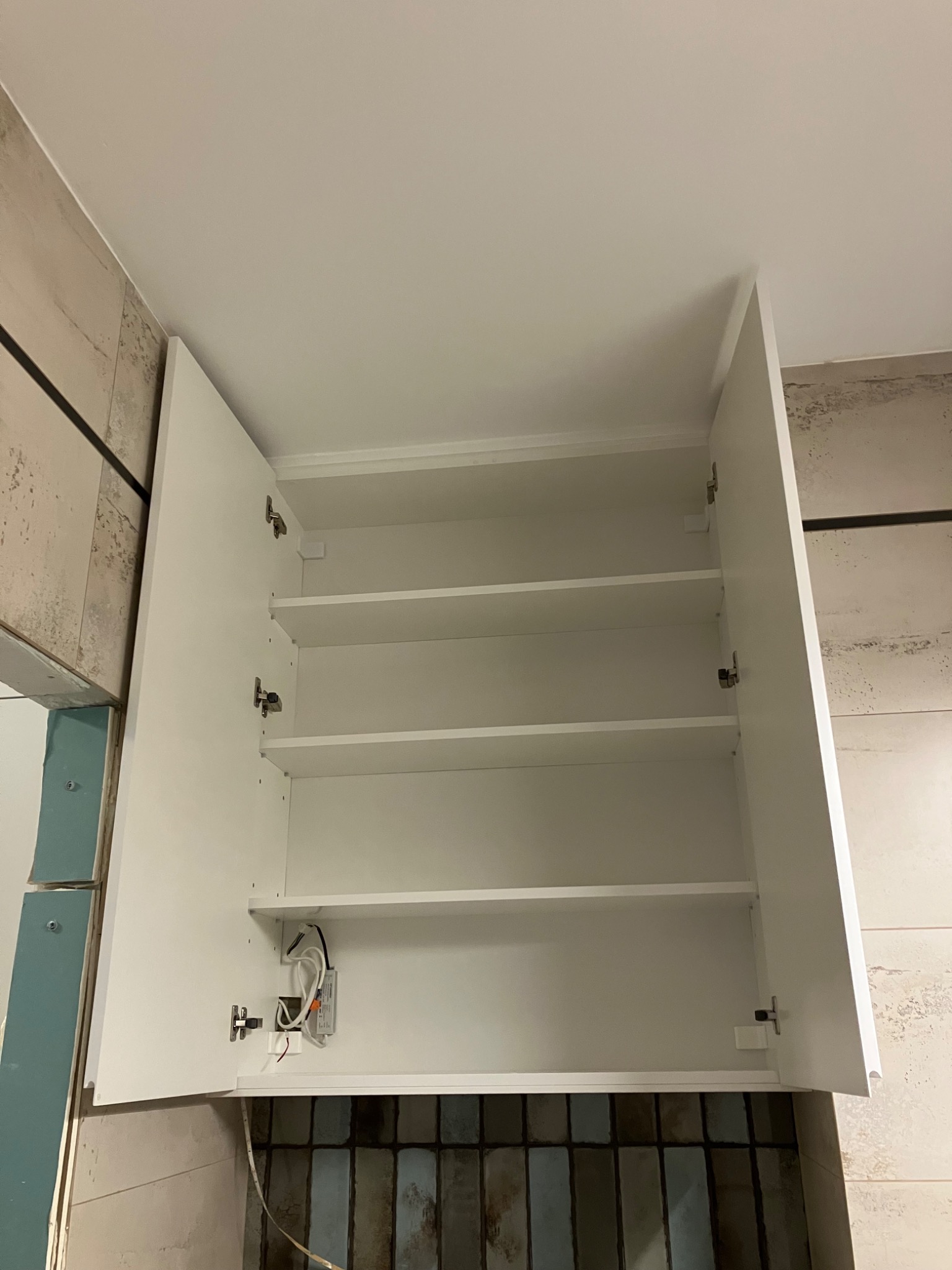 Biała szafka kuchenna wisząca z otwartymi drzwiczkami, widoczne półki i oświetlenie LED. Montaż szafki w trakcie wykończenia kuchni, widoczne płytki i surowa ściana.