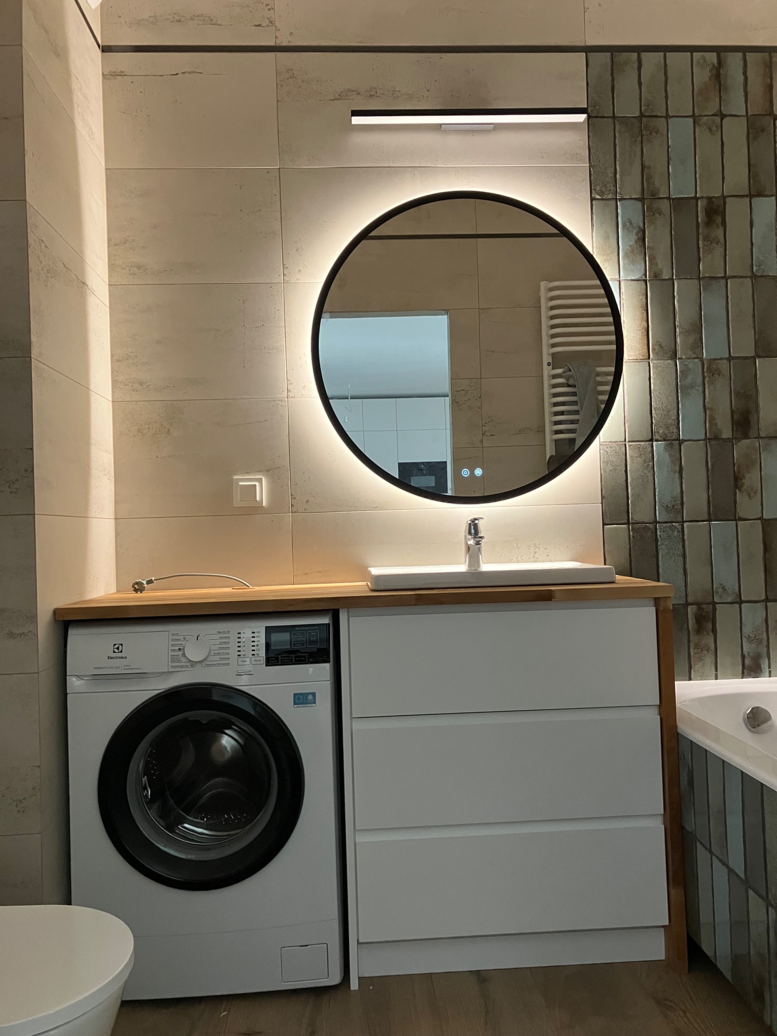 Nowoczesna łazienka z pralką zabudowaną pod blatem z umywalką i okrągłym lustrem z podświetleniem LED. Minimalistyczny design, płytki w odcieniach beżu i szarości.