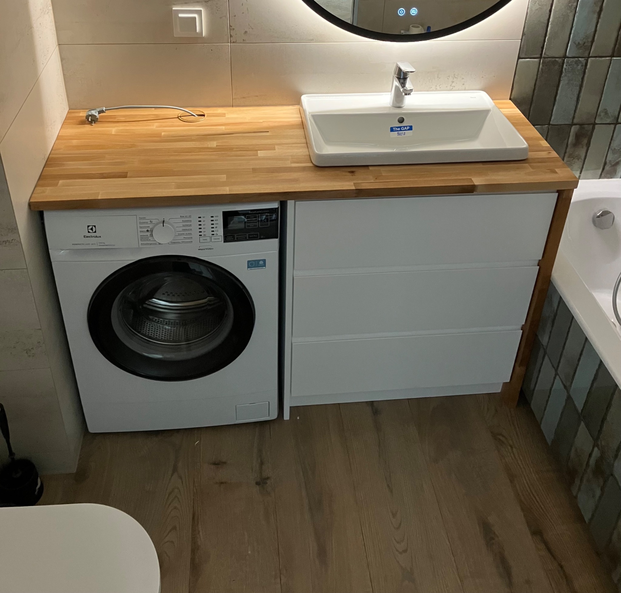 Nowoczesna łazienka z drewnianym blatem, białą umywalką Roca i pralką Electrolux w zabudowie. Minimalistyczny design, ciepłe oświetlenie i drewniana podłoga.