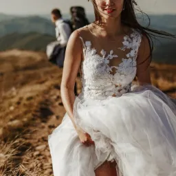 Panna młoda w koronkowej sukni ślubnej z tiulowym dołem, trzymająca suknię w dłoni, spaceruje po górskim szlaku; w tle mężczyzna z plecakiem i rozległy krajobraz górski, autor: Tomasz Walowski.