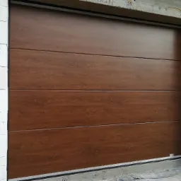 Segmentowa brama garażowa w kolorze drewna, zamontowana w surowym otworze z białych bloczków betonowych.