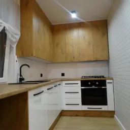 Nowoczesna kuchnia z białymi szafkami dolnymi, drewnopodobnymi szafkami górnymi, czarną armaturą i piekarnikiem, oraz ścianą wykończoną imitacją białej cegły.