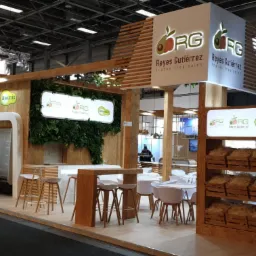 Stoisko targowe z drewnianą konstrukcją, logo firmy z awokado i tropikalnymi owocami, drewniane kraty, zielona ściana i minimalistyczne meble.