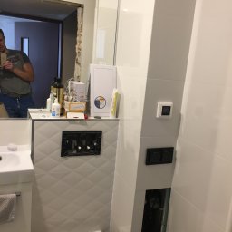 Eneo2012 - Biała łazienka w trakcie remontu: widoczna toaleta, umywalka z szafką, płytki z wzorem rombów, instalacja podtynkowa i mężczyzna robiący zdjęcie w lustrze.