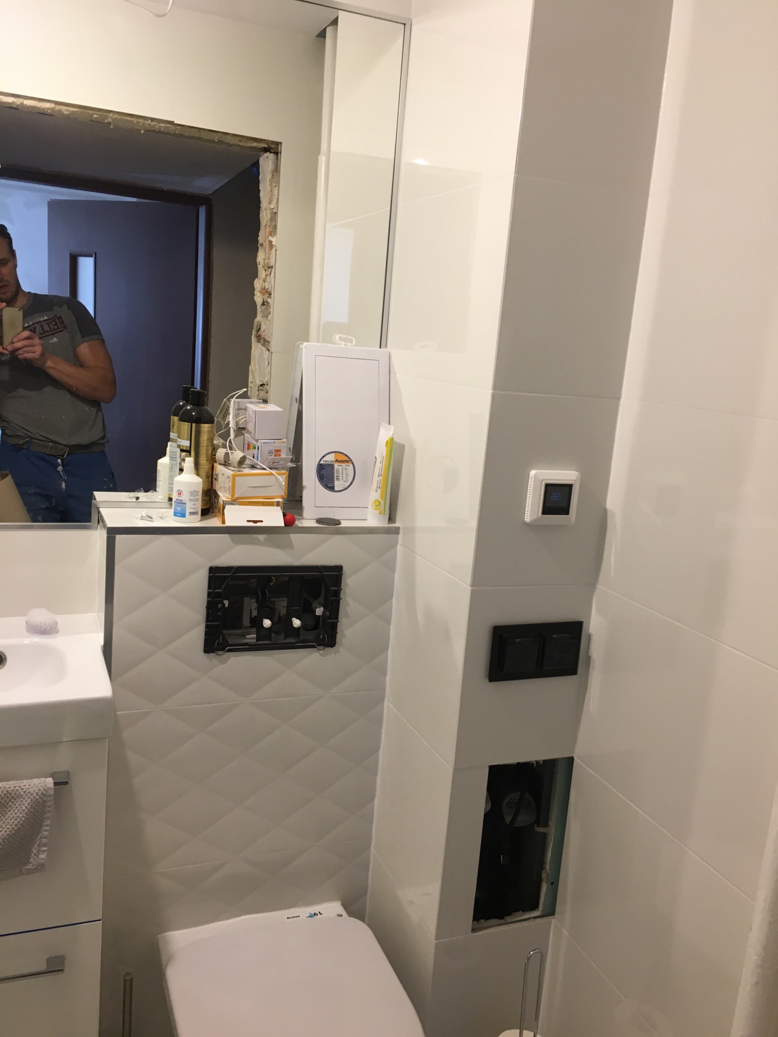 Biała łazienka w trakcie remontu: widoczna toaleta, umywalka z szafką, płytki z wzorem rombów, instalacja podtynkowa i mężczyzna robiący zdjęcie w lustrze.