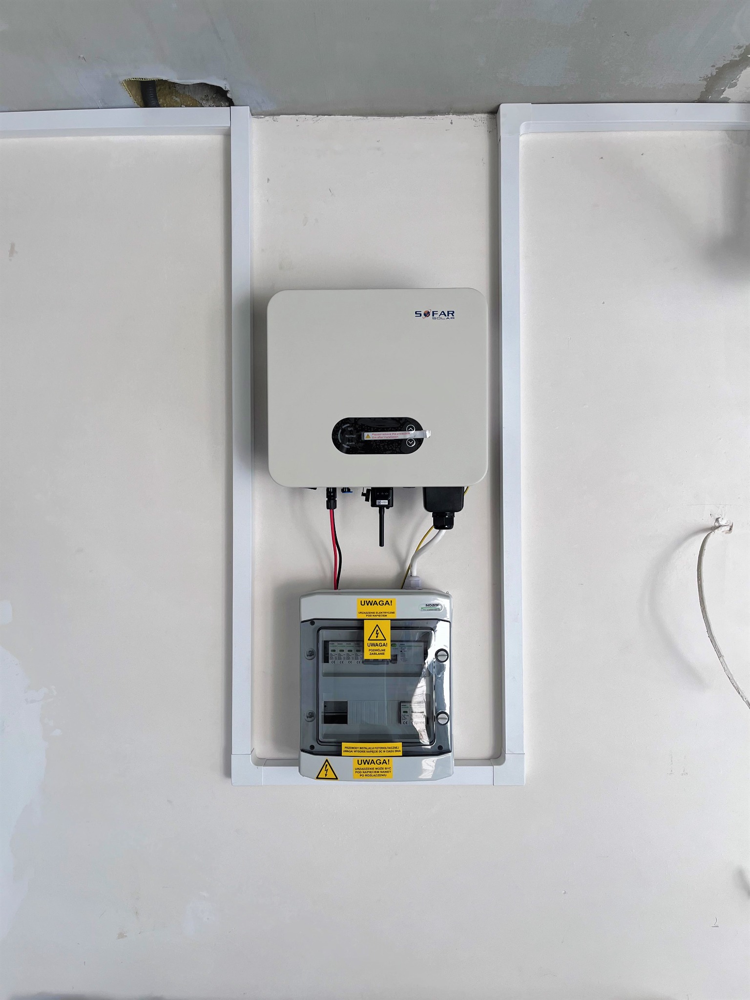 Biały inwerter SOFAR oraz szara skrzynka zabezpieczeń fotowoltaicznych zamontowane na białej ścianie w białej ramie. Widoczne kable i przewody. Niedokończone wykończenie sufitu w tle.