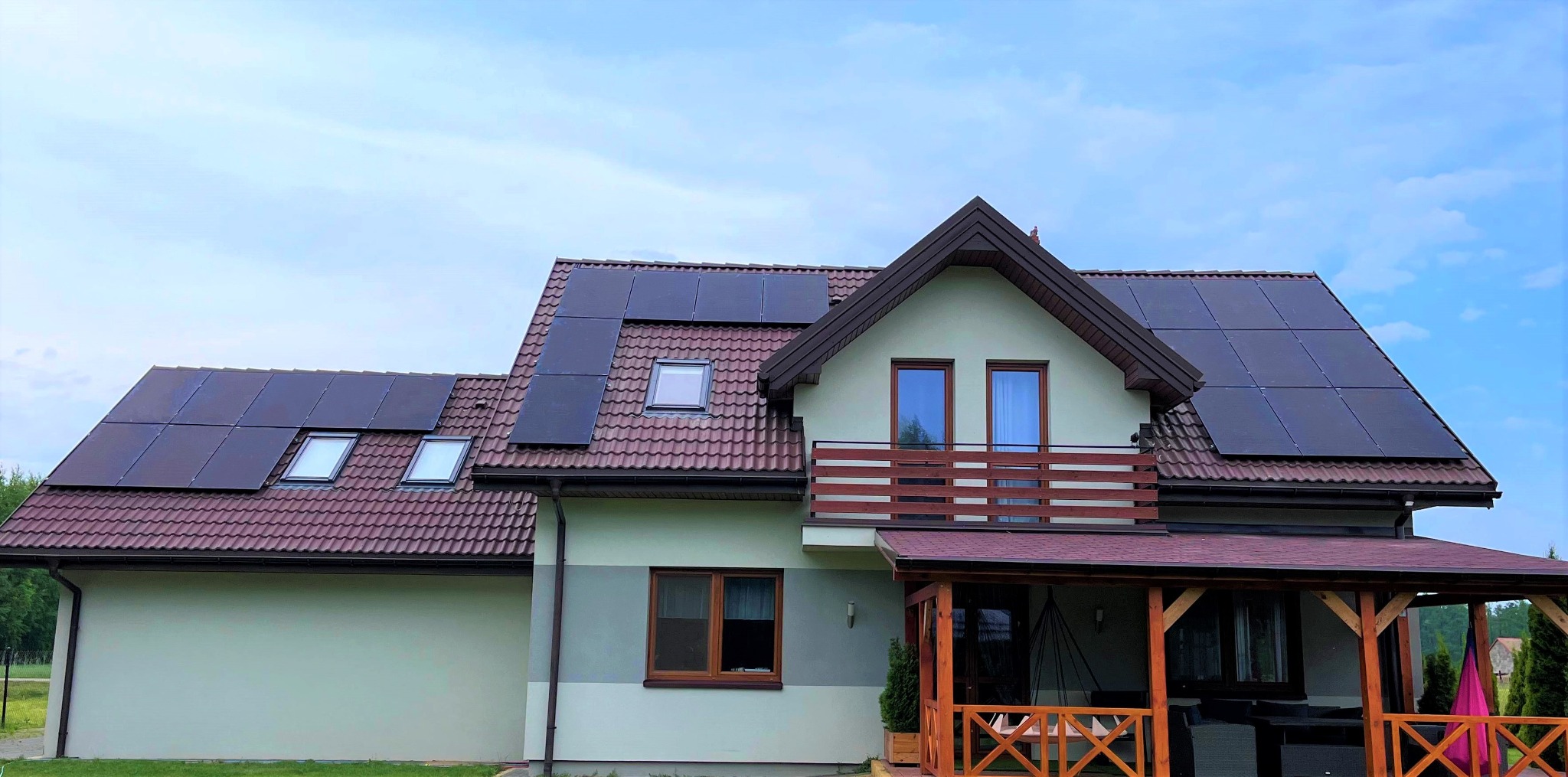 Dom jednorodzinny z panelami słonecznymi zamontowanymi na dachu z dachówką ceramiczną, widoczne okna dachowe i balkon.