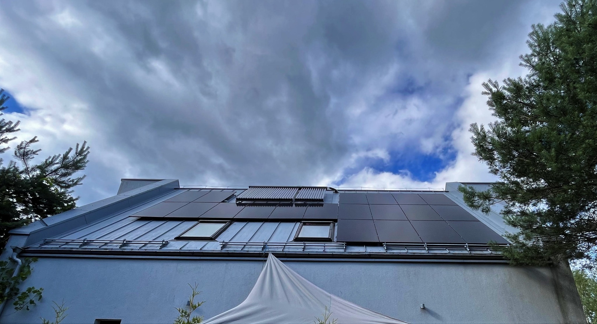 Czarny panel fotowoltaiczny i kolektor słoneczny na szarym blaszanym dachu domu jednorodzinnego, widok z dołu na tle pochmurnego nieba.