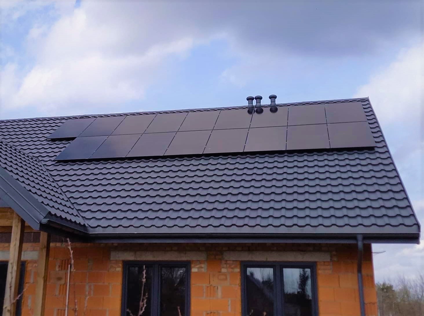 Panele słoneczne zamontowane na dachu domu w trakcie budowy, widoczne ceglane ściany i ciemne okna.