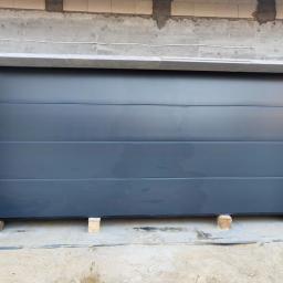 Hastam - Nowa, segmentowa brama garażowa w kolorze antracytowym, z widocznym logo producenta, tymczasowo podparta drewnianymi klockami w trakcie montażu w surowym otworze garażowym.