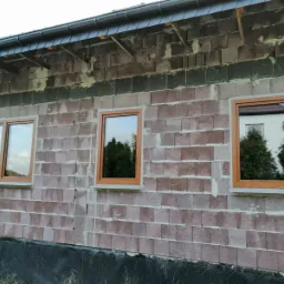 Montaż okien z brązowymi ramami w surowym murze budynku w Myszkowie, widoczne refleksy otoczenia w szybach.