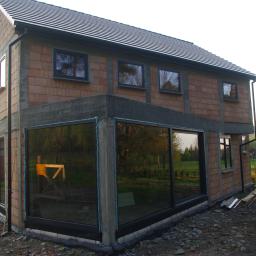 Hastam - Surowy budynek w trakcie budowy z zamontowanymi oknami z czarnymi ramami, widoczne cegły i szare elementy betonowe, w oknach odbija się zieleń drzew.