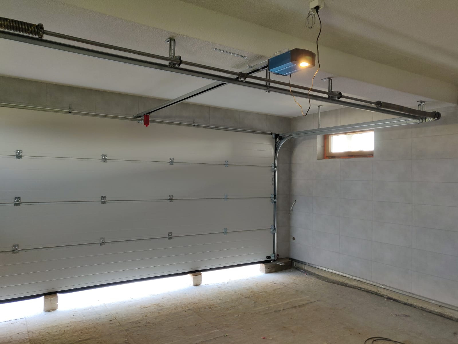 Częściowo otwarta segmentowa brama garażowa z napędem elektrycznym zamontowanym pod sufitem, widoczne sprężyny i szyny prowadzące.