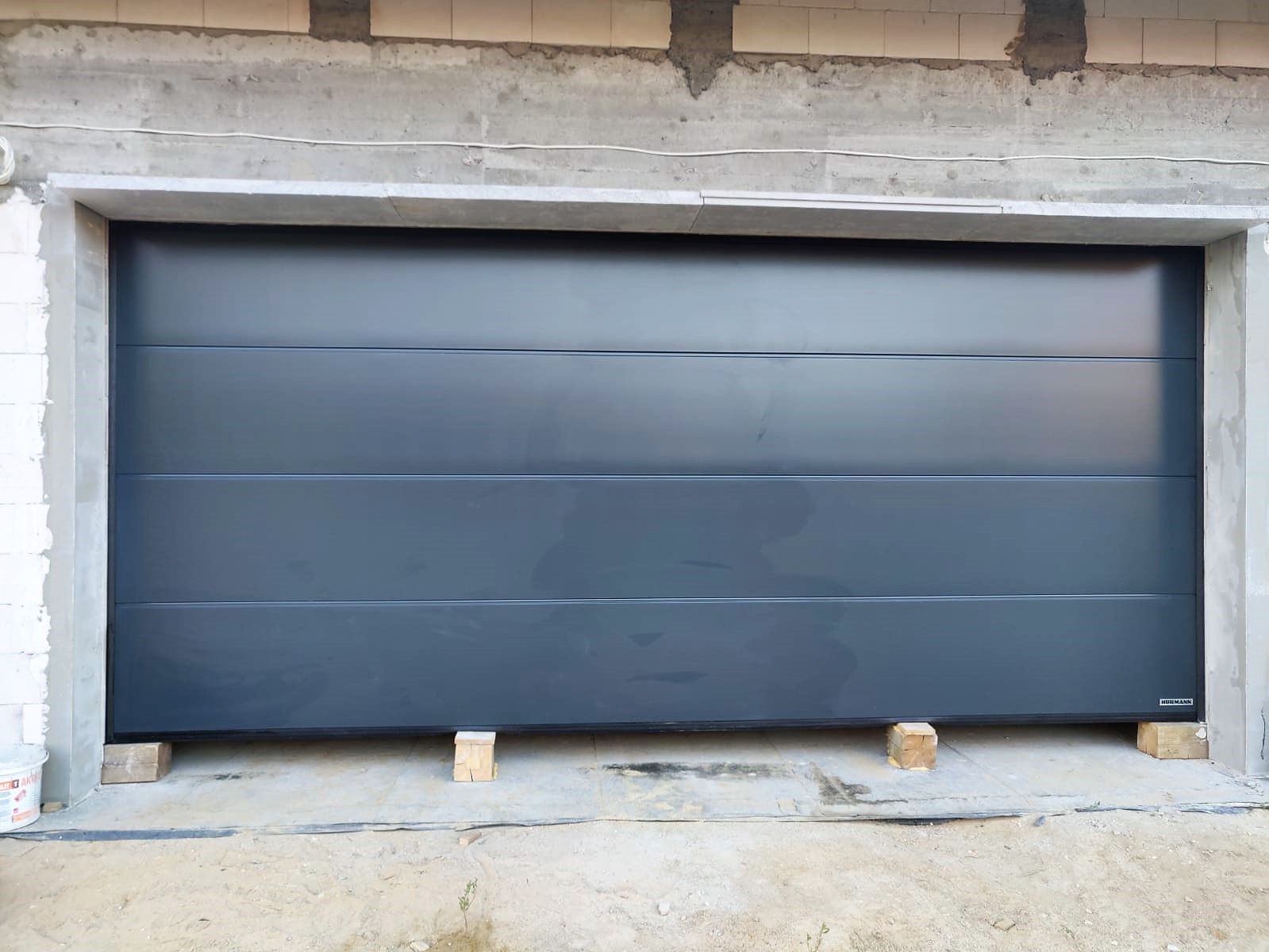 Nowa, segmentowa brama garażowa w kolorze antracytowym, z widocznym logo producenta, tymczasowo podparta drewnianymi klockami w trakcie montażu w surowym otworze garażowym.