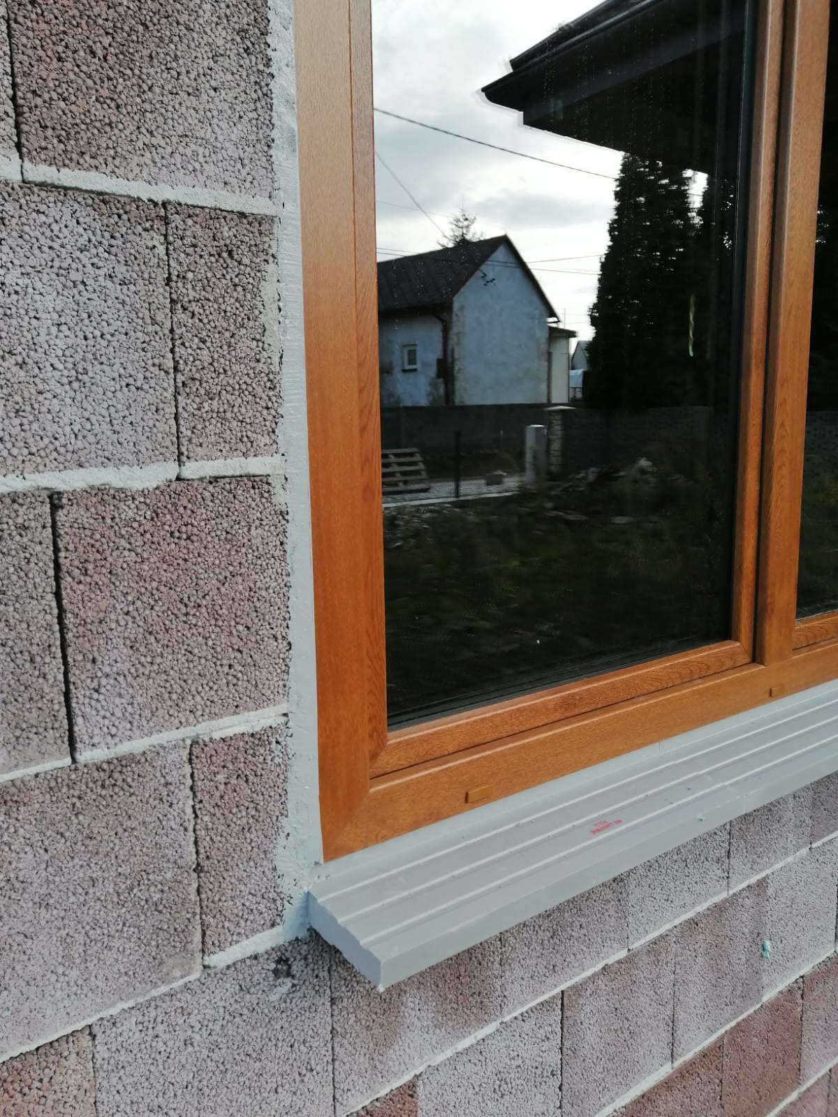 Nowe okno z drewnianą ramą zamontowane w ścianie z bloczków betonowych, widoczny parapet zewnętrzny i odblask domu w szybie.