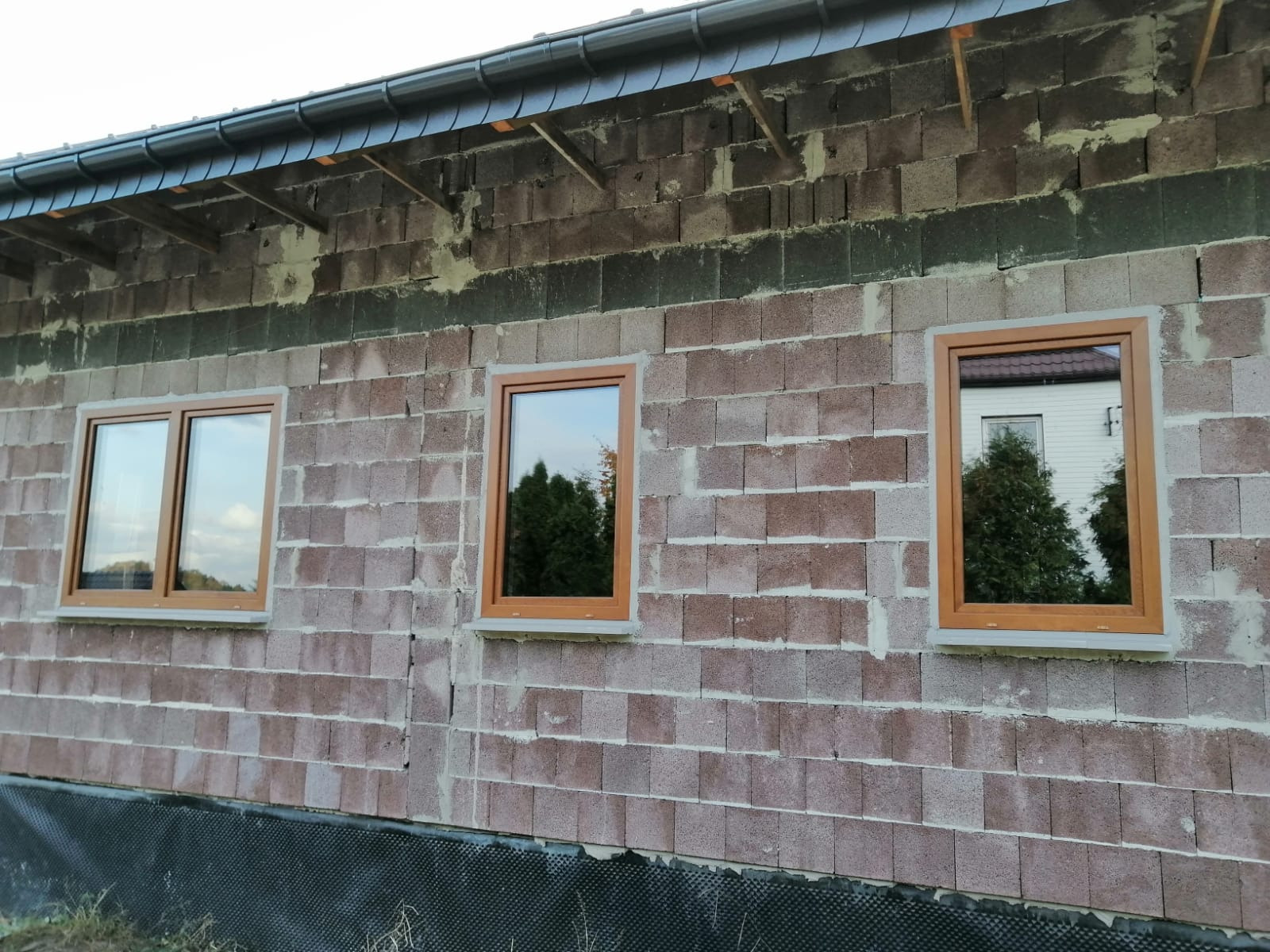 Montaż okien z brązowymi ramami w surowym murze budynku w Myszkowie, widoczne refleksy otoczenia w szybach.