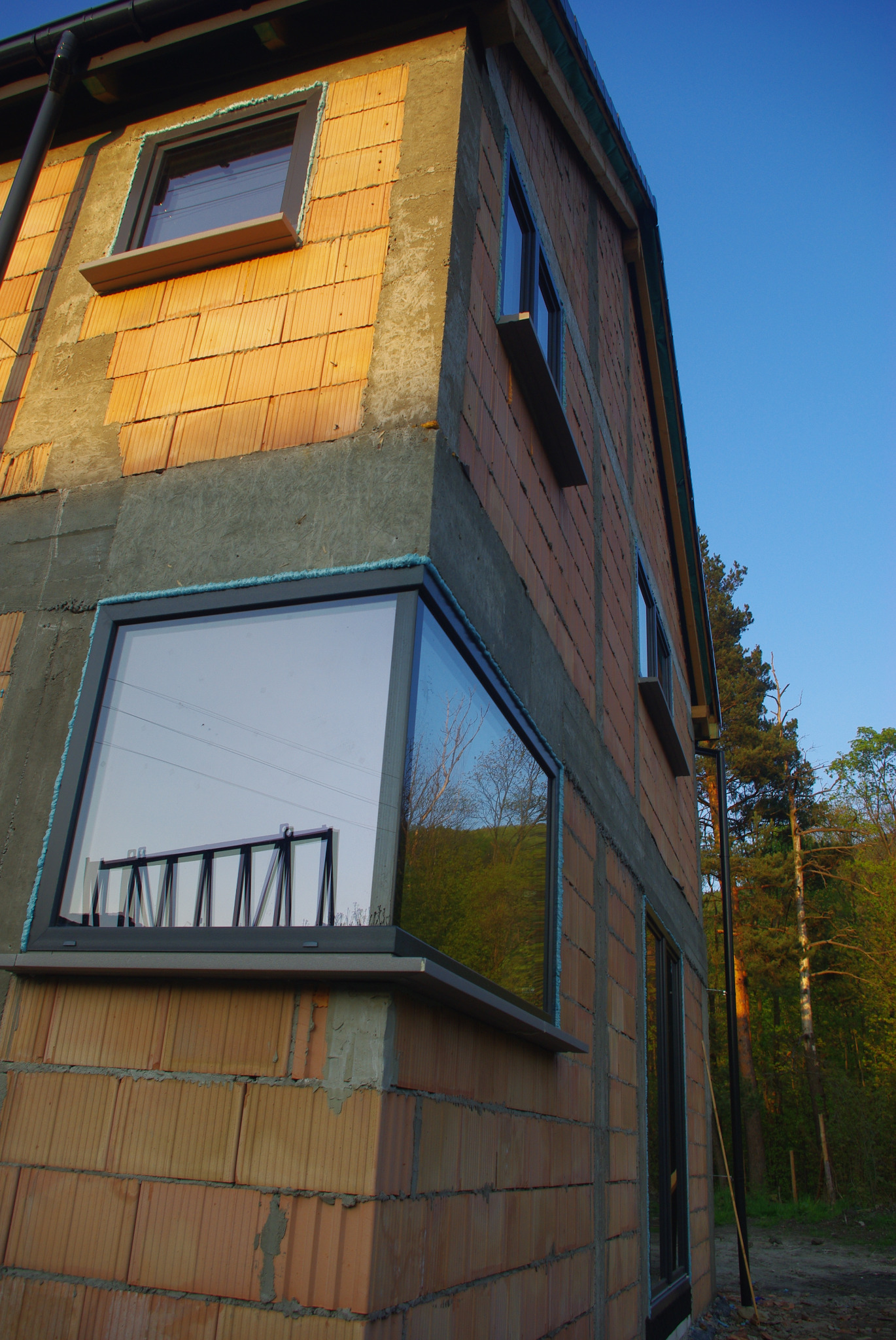 Niedokończony budynek z zamontowanymi oknami o szarych ramach, widoczna cegła i pianka montażowa, odbicie krajobrazu w szybach, błękitne niebo w tle.