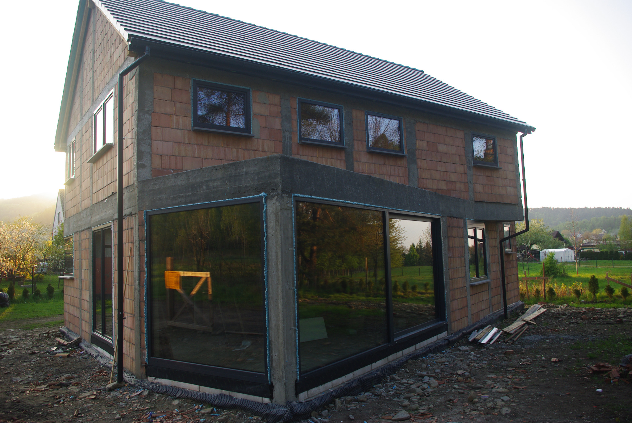 Surowy budynek w trakcie budowy z zamontowanymi oknami z czarnymi ramami, widoczne cegły i szare elementy betonowe, w oknach odbija się zieleń drzew.