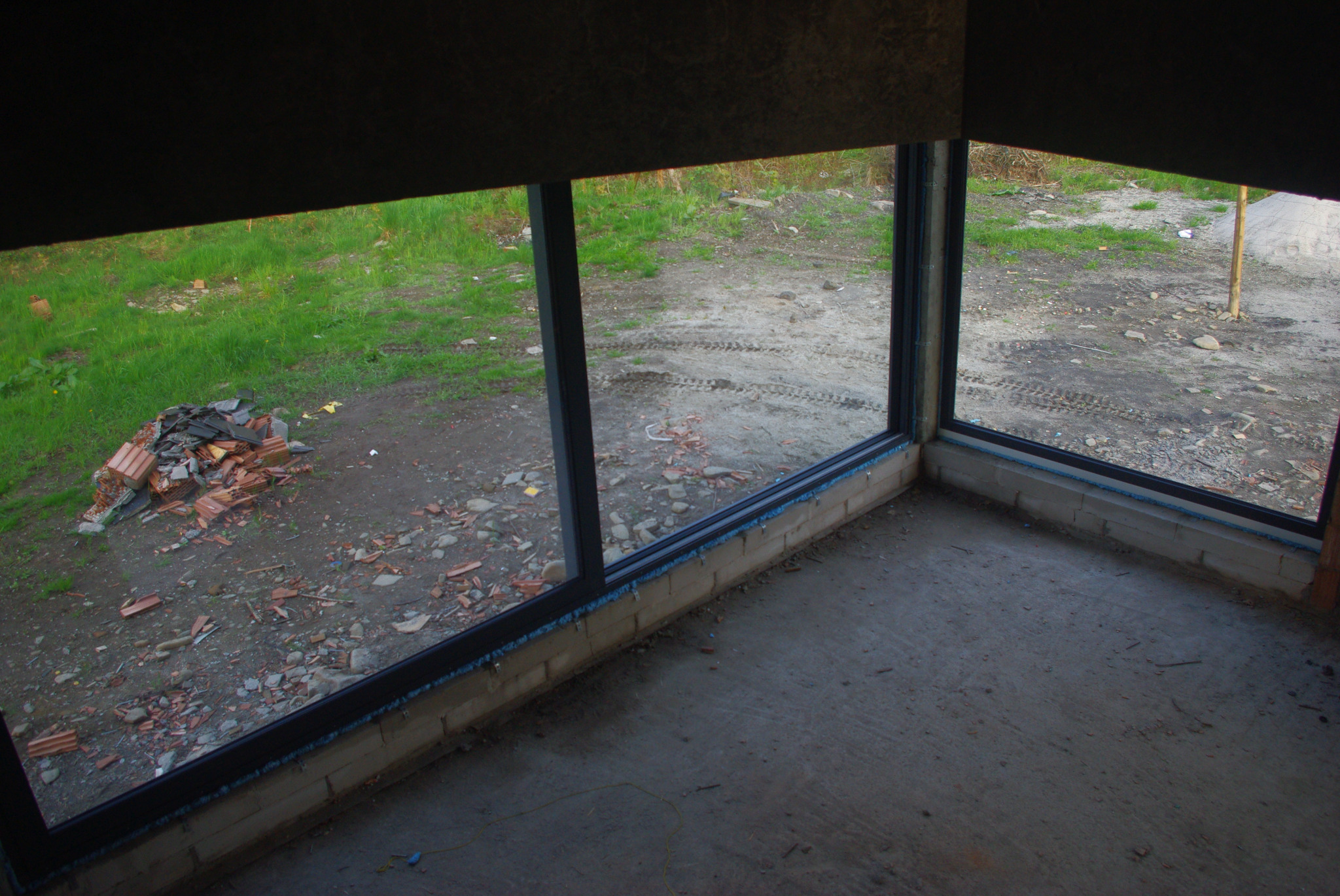 Surowy stan wnętrza z zamontowanymi oknami z czarnymi ramami; widok na zewnątrz na gruz i zieloną trawę.