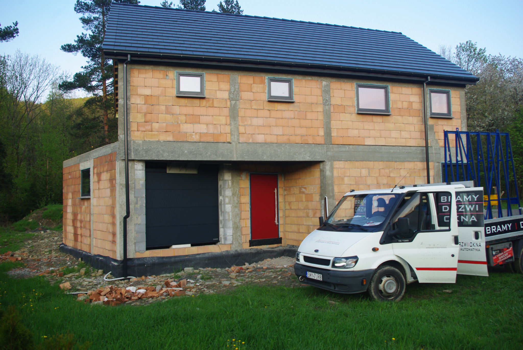 Niedokończony dom z cegły z zamontowanymi oknami i bramą garażową, czerwone drzwi wejściowe, na trawniku stoi biały samochód dostawczy z reklamą okien i drzwi.