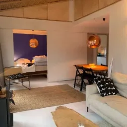 Jasne wnętrze w stylu skandynawskim z drewnianym sufitem, widok na sypialnię z niebieską ścianą, lampy wiszące w kolorze miedzi, stolik kawowy z blatem z surowego drewna.