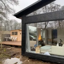 Nowoczesny dom z czarną drewnianą elewacją i dużymi oknami, widok zewnętrzny na taras z domkiem letniskowym i meblami ogrodowymi, odbicie wnętrza domu w szybach.