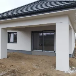 Nowoczesny dom jednorodzinny w trakcie wykończenia, widoczne czarne okna i przesuwne drzwi tarasowe, żółty przewód na elewacji, teren wokół domu w trakcie zagospodarowania.