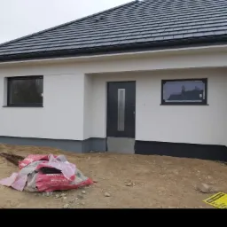 Nowoczesny, parterowy dom jednorodzinny w trakcie wykończenia, z szarymi oknami i drzwiami, widoczne materiały budowlane na niezagospodarowanym terenie.