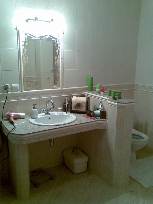 Umywalka w łazience z blatem z jasnego kamienia, podwieszana szafka i lustro z dekoracyjnymi elementami, widoczne płytki ścienne i podłogowe w odcieniach beżu.