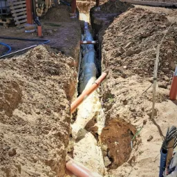 Głęboki wykop z rurami zabezpieczonymi pianką izolacyjną, widok z góry ukazuje nierówności terenu i drewnianą konstrukcję w tle.