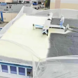 Ocieplanie dachu pianką PUR w trakcie realizacji, widoczni dwaj pracownicy w kombinezonach ochronnych, jeden aplikuje piankę, drugi nadzoruje, część dachu już pokryta jasną pianką, reszta w kolorze...