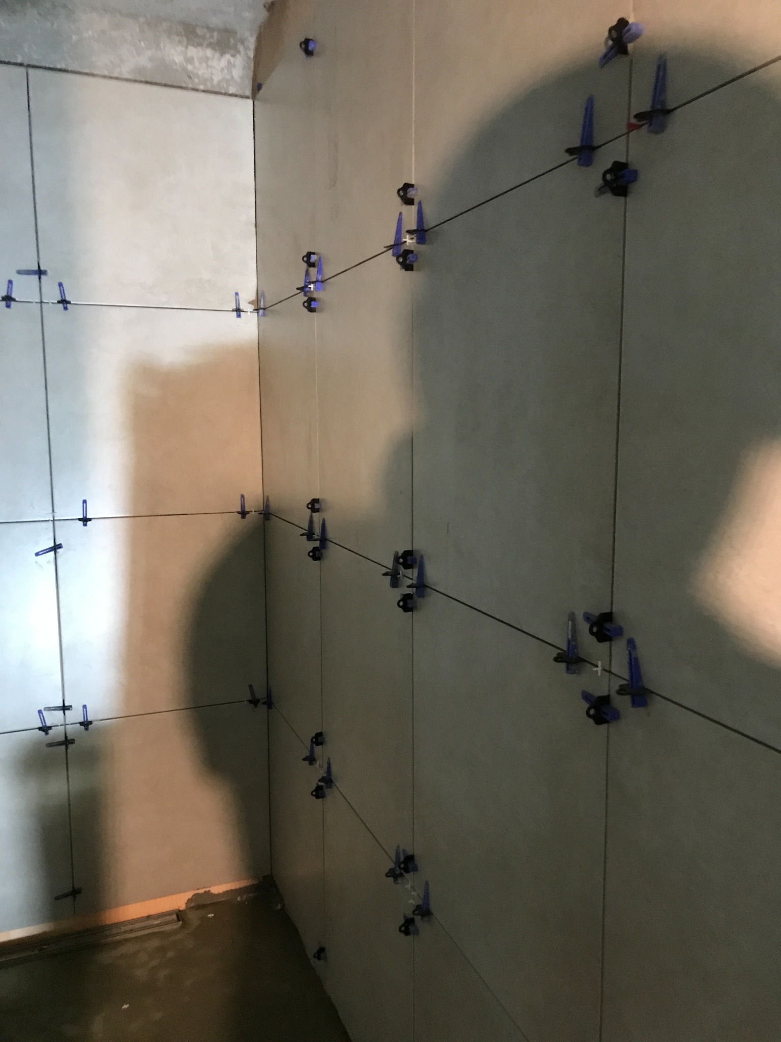 Świeżo ułożone, jasnoszare płytki ścienne w rogu łazienki, stabilizowane niebieskimi klipsami i czarnymi klinami dystansowymi, oczekujące na fugowanie.