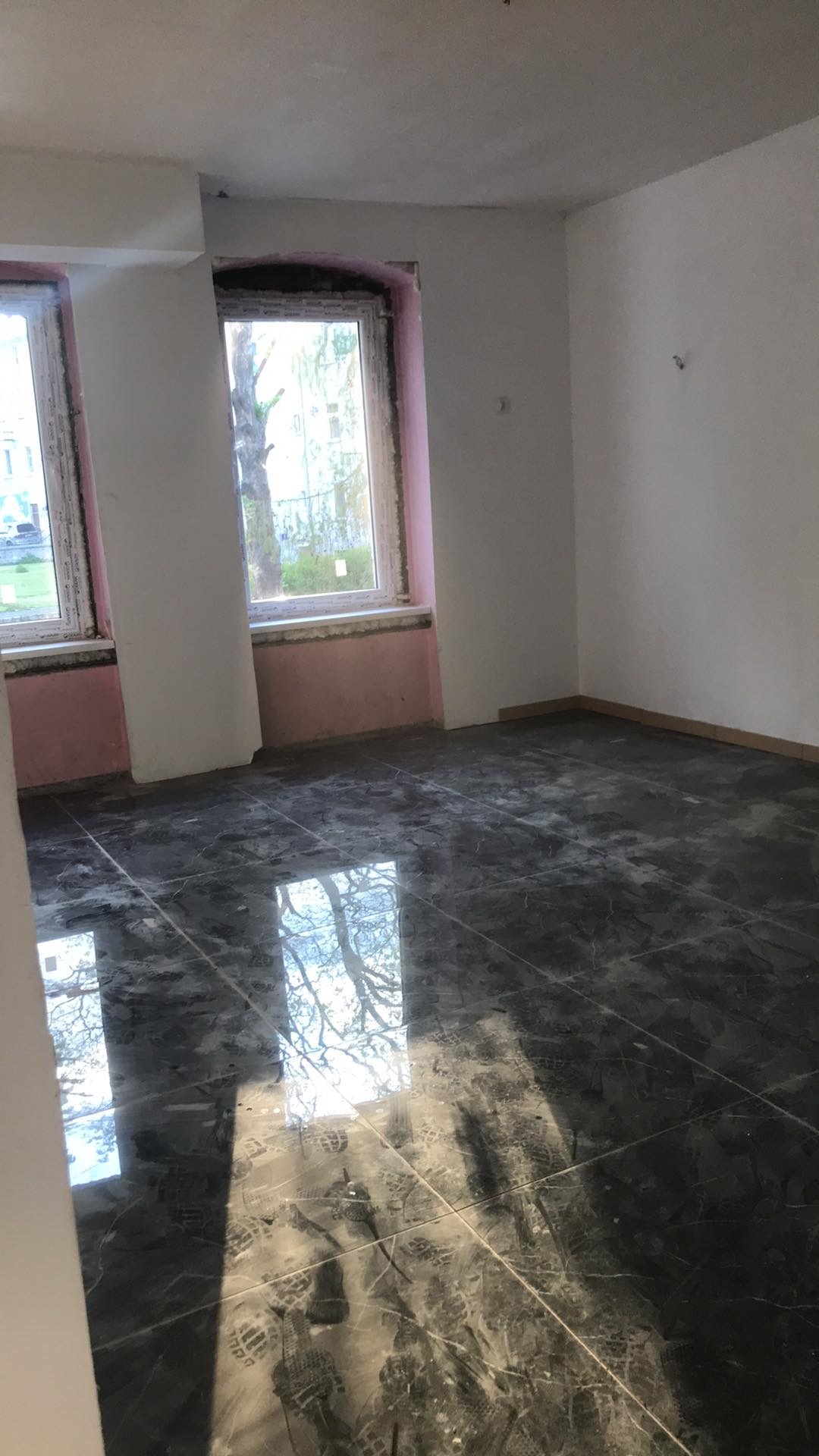 Wnętrze pokoju z nową, błyszczącą, ciemną podłogą z płytek imitujących marmur, odbijającą światło i widok drzewa za oknem; ściany częściowo przygotowane do malowania, z widocznymi śladami po...