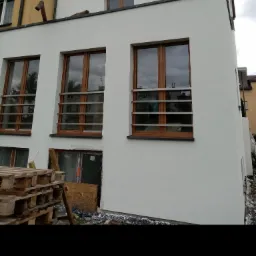 Nowoczesny budynek w Lublinie z białymi ścianami i drewnianymi oknami z metalowymi barierkami, widoczne palety i materiały budowlane wskazujące na trwające prace wykończeniowe.