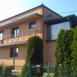 Usługi remontowo budowlane Paweł Magda - Dwupiętrowy dom w odcieniach beżu z balkonem zabezpieczonym metalową balustradą, widoczny zza zielonego żywopłotu i siatki ogrodzeniowej.