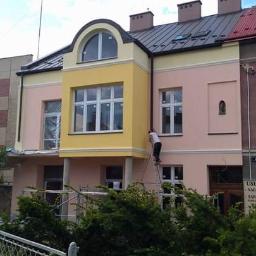 Usługi remontowo budowlane Paweł Magda - Pracownik na drabinie maluje elewację budynku w pastelowych kolorach, widoczny fragment rusztowania i tablica z napisem 'Usługi - Nagrobki - Tablice'.