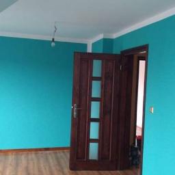 Usługi remontowo budowlane Paweł Magda - Pokój po odświeżeniu ścian w turkusowym odcieniu, z drewnianą podłogą i drzwiami z przeszkleniami, widoczna surowa żarówka.