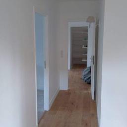 Usługi remontowo budowlane Paweł Magda - Korytarz z drewnianą podłogą po remoncie, świeżo pomalowane białe ściany i ościeżnice, widok na otwarte drzwi do garderoby z półkami.