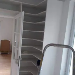 Wnętrze z narożną, białą szafą wnękową z otwartymi półkami i przeszklonymi drzwiami, widoczna drabina malarska, jasna podłoga i okno.