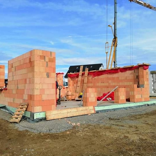 Wznoszenie ścian domu z czerwonej cegły ceramicznej Porotherm, widoczny dźwig budowlany i drewniane schody tymczasowe.