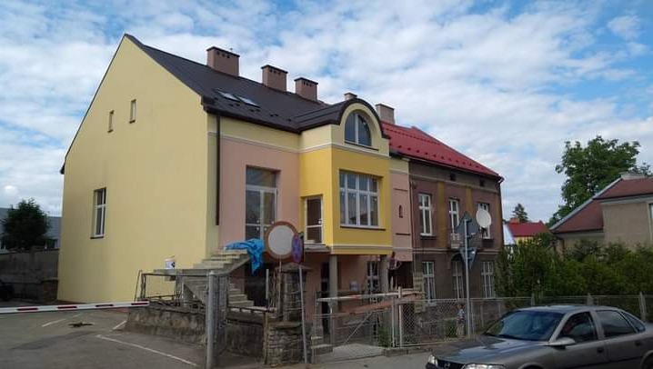 Odnowiona fasada kamienicy w pastelowych kolorach, z żółtą elewacją po lewej stronie i różową po prawej, widoczne schody wejściowe i zaparkowany samochód na pierwszym planie.