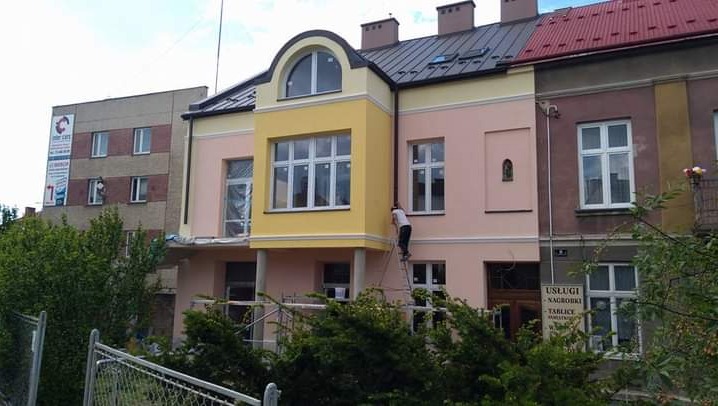 Pracownik na drabinie maluje elewację budynku w pastelowych kolorach, widoczny fragment rusztowania i tablica z napisem 'Usługi - Nagrobki - Tablice'.