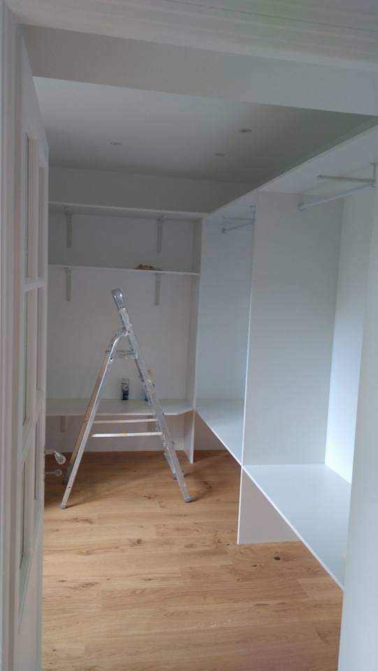 Wnętrze garderoby w trakcie wykańczania: białe półki i wieszaki, drewniana podłoga, aluminiowa drabina oparta o ścianę, widok zza otwartych drzwi.